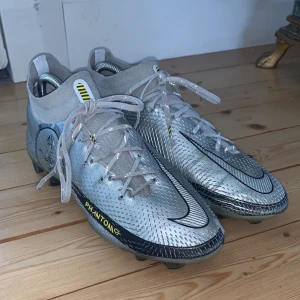 Nike Phantom GT2 Elite fotbollsskor - Säljer ett par Nike Phantom GT2 Elite fotbollsskor i metallic silver med svarta och gula detaljer. Dobbar för gräsplan. Skorna är änvända en del. Smutsiga på några ställen, söndrig på ett ställe. Men annars mycket kvar att ge. Skick 7/10. 
