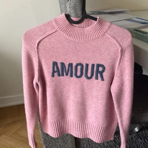 Rosa stickad tröja med text från Zadig & Voltaire - Säljer en rosa  tröja från Zadig & Voltaire med blå texten 'AMOUR' framtill. Tröjan har ribbade muddar vid ärmslut, hals och nederkant samt lång ärm. Perfekt för dig som gillar en mysig och trendig look.