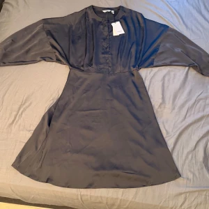 Grå satinklänning från b.young - Säljer en grå satinklänning från b.young med knappar framtill och lång ärm. Klänningen har en lätt utställd kjol och markerad midja för en smickrande siluett. Den är helt oanvänd och skriv gärna vid frågor om måtten.