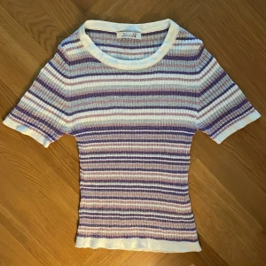 Randig ribbad topp från Design by Si - Säljer en snygg ribbad topp från Design by Si i storlek XS/S. Tröjan har korta ärmar och är randig i vitt, lila och rosa med inslag av glittrigt material. 