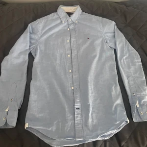 Ljusblå skjorta från Tommy Hilfiger - Snygg ljusblå skjorta från Tommy Hilfiger med klassisk button-down krage och små broderade loggan på bröstet. Skjortan har vita knappar och lång ärm, perfekt till en stilren look. Skjortan är väl omhändertagen. 