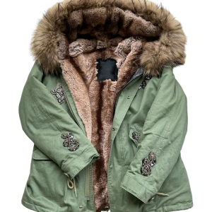 Grön parkajacka strö M med tvättbjörn päls och broderier - Parkajacka i militärgrönt  lyxig  och stor tvättbjörn pälskantad huva. Snygga broderade detaljer på ärmar och bröst, samt snörning i midjan. Jackan har dragkedja och knappar framtill och är perfekt för kalla dagar.Nypris va 650 euro, har aldrig burit jackan, den är helt ny