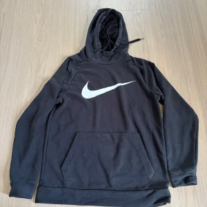 Svart Nike hoodie Dri-Fit S - Svart hoodie från Nike med stor vit Swoosh-logga på bröstet. Tröjan har huva med snörning, känguruficka och är tillverkad i Dri-Fit polyester som andas. Perfekt för träning eller chill, med lång ärm och klassisk passform.