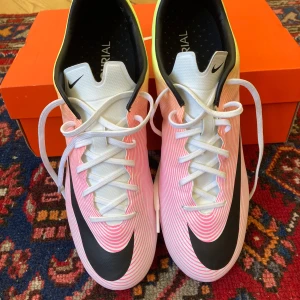Nike Mercurial Vapor 2 fg  - Säljer ett par Nike Mercurial fotbollsskor med snyggt randigt mönster i vitt, rosa och gult. Skorna har svarta detaljer, stora Nike-loggor och gula dobbar. Ovandelen är i syntetmaterial och de har klassisk snörning. Perfekta för dig som vill sticka ut på planen.