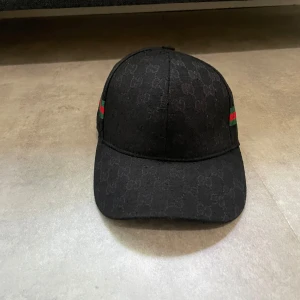 Svart keps från Gucci med GG-mönster - Svart keps från Gucci med diskret GG-monogram över hela ytan och klassiska gröna och röda ränder på sidorna. Justerbar rem baktill för perfekt passform. Snygg och stilren accessoar som lyfter din outfit.