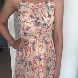 Blommig ljus kortklänning med volang - Superfin ljus kortklänning med blommigt mönster i rosa, lila och blå toner. Klänningen har volangdetaljer framtill, är ärmlös och har en lätt, flowy passform. Perfekt för sommarens alla tillfällen!