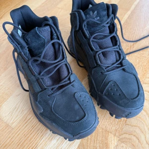 Svarta adidas vinter/höst sneakers med grov sula - Svarta adidas sneakers med högt skaft och grov, mönstrad yttersula för extra grepp. Skorna har snörning, mockadetaljer och klassiska tre ränder på sidan. Ovandelen är en mix av mocka och textil, vilket ger en cool och sportig vibe. Varma och perfekta för höst och vinter. Köpt till herr men funkar unisex. Använda en gång. 