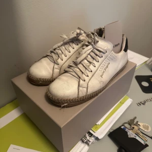 Palm Angels vita sneakers med svarta detaljer - Palm Angels sneakers i vitt skinn med svarta mockadetaljer på hälen och grov sula med repdetalj. Klassisk låg modell med snörning och rund tå. Märkeslogga på sidan och mönstrad gummisula undertill. Perfekta för dig som gillar streetstyle. Möts i Stockholm mvh