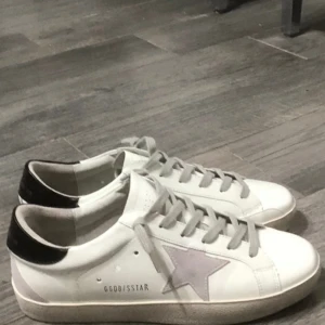 Golden Goose vita sneakers med stjärna - Snygga vita sneakers från Golden Goose med grå mockastjärna på sidan, grå snörning och svart detalj på hälen. Skorna har en platt sula och GGDB/SSTAR-tryck på sidan. Perfekta för en cool och avslappnad stil.