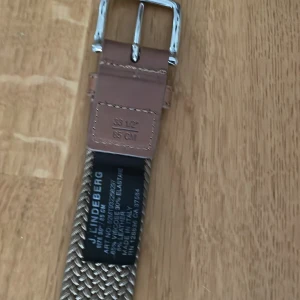 Beige flätat bälte från J.Lindeberg - Snyggt flätat bälte från J.Lindeberg i beige med detaljer i brunt skinn och silvrigt spänne. Bältet har en stilren design med vävd struktur och präglad logga på läderdetaljen. Perfekt accessoar för att lyfta din outfit.