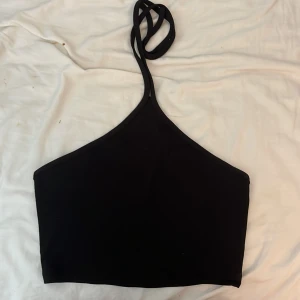 Svart oneshoulder topp med knyt - Snygg svart topp med oneshoulder-design och smal axelrem som knyts i nacken. Croppad modell som passar perfekt till höga jeans eller kjol. Enkel och stilren look som är lätt att matcha.