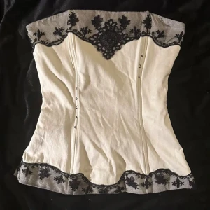 Vit och svart korsett från Vila XS - Supersnygg vit korsett från Vila i storlek XS med svarta spetsdetaljer och broderade blommor längs kanterna. Toppen är axelbandslös och har en elegant, figurnära passform. 