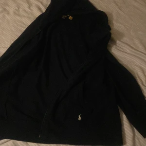 Svart hoodie från Polo Ralph Lauren - Svart hoodie från Polo Ralph Lauren med klassisk broderad logga på bröstet. Tröjan har huva, dragkedja framtill och långa ärmar. Tillverkad i mjuk bomull som känns skön mot huden. Perfekt för en chill och stilren look. Äkta🔥