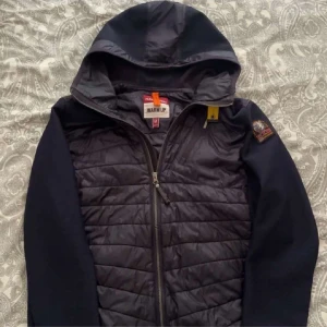 Svart pufferjacka från Parajumpers - Snygg svart pufferjacka från Parajumpers med huva och coolt PJS-märke på ärmen. Jackan har quiltad front, dragkedja och detaljer i syntetmaterial. Perfekt för dig som vill ha en sportig och urban look.