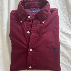 Vinröd skjorta från Ralph Lauren - Vinröd skjorta från Ralph Lauren med klassisk krage och vita knappar. Broderad logga på bröstet och tillverkad i mjuk bomull. Perfekt för dig som gillar stilrena plagg. Storlek S