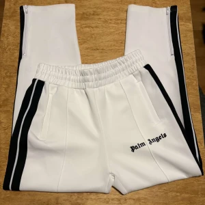 Palm Angels vita trackpants med svarta ränder - Palm Angels vita trackpants med svarta sidoränder och broderad logga på benet. Byxorna har elastisk midja med snörning, raka ben med dragkedja vid anklarna och är gjorda i ett mjukt syntetmaterial. Perfekta för en clean streetwear-look.