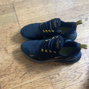 Nike Air 270 svarta sneakers med gula detaljer - Nike Air 270 sneakers i svart med snygga gula detaljer och loggor. Ovandelen är i ventilerande mesh och skorna har en tydlig Air-sula baktill. Klassisk snörning och sportig vibe, perfekt för dig som gillar streetstyle och komfort. Har haft dem cirka 3 månader men tyvär blev dem för små.