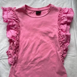 Rosa volangtopp från Gina Tricot - Supersöt rosa blus från Gina Tricot med volangdetaljer längs axlarna. Toppen har rund halsringning och är ärmlös, perfekt för dig som gillar en lekfull och trendig stil. Tillverkad i mjuk bomull som känns skön mot huden.Säljer för att den inte används💗Skriv för funderingar eller fler bilder🍓