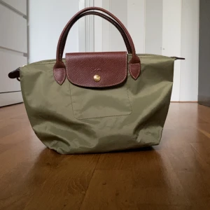 Grön Longchamp Le Pliage handväska - Säljer en klassisk grön Longchamp Le Pliage handväska med bruna skinnhandtag och lock. Väskan har guldfärgade detaljer och stängs med dragkedja och tryckknapp. Perfekt storlek för vardagens essentials och enkel att bära i handen eller på armen.