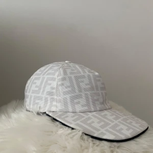 Vit Fendi keps med FF-mönster - Snygg vit fendi keps med ikoniskt FF-logomönster över hela kepsen. Klassisk böjd skärm och justerbar rem baktill. Tillverkad i ett tåligt tyg med svart insida för extra kontrast. Perfekt för dig som vill sticka ut med en lyxig accessoar.