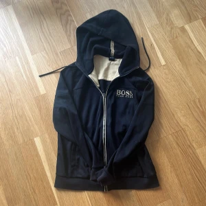 Svart hoodie från Hugo Boss - Svart hoodie från Hugo Boss med broderad logga på bröstet och dragkedja framtill. Insidan av huvan är beige och huvan har snörning. Tröjan är långärmad och har ribbade muddar. Perfekt för en chill och stilren look.
