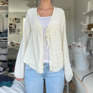 Krämvit omlottkofta med knyt - Supersöt krämvit kofta med omlottkänsla och knyt framtill. Lätt strukturerat tyg som ger en chill och luftig vibe. Långärmad och perfekt att slänga över ett linne eller topp. Passar till både jeans och kjol för en effortless look.