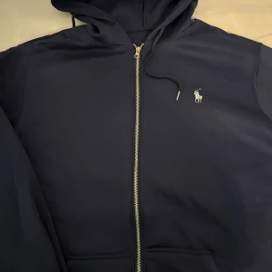 Marinblå hoodie från Ralph Lauren - Snygg marinblå hoodie från Ralph Lauren med dragkedja och klassisk vit logga broderad på bröstet. Tröjan har huva med snörning och två fickor framtill. Perfekt för en chill och stilren look.
