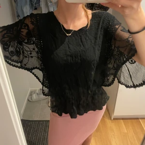 Svart blus med spets och vida ärmar - Säljer en svart blus med transparenta, vida ärmar i spets och broderade blommor. Toppen har en lös passform och vacker volangkant nertill. Perfekt för dig som gillar unika detaljer och en romantisk vibe.