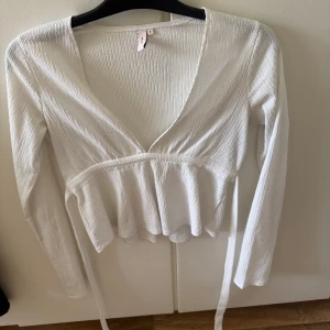 Vit omlottopp från Nelly - Supersöt vit omlottopp från Nelly med lång ärm och knyt i midjan. Toppen har v-ringning och lätt rynkad struktur i tyget som ger en snygg och trendig look. Perfekt att styla med jeans eller kjol för en fräsch vibe.