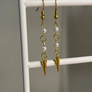 Guldiga örhängen med pärlor och spikes - Snygga hängande örhängen i guldton med små vita pärlor och coola konformade spikes längst ner. Kedjedetaljerna ger en edgy vibe och pärlorna gör dem extra stilrena. Perfekta för dig som vill sticka ut lite extra. Handgjorda