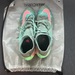 Nike Mercurial fotbollsskor mint/grön - Nike Mercurial fotbollsskor i mintgrönt med neonrosa detaljer och svart/grön sula. Skorna har snörning, högre strumpliknande skaft och stor Nike-logga på sidan. Ovandelen är i syntetmaterial och designen är riktigt sportig och modern.