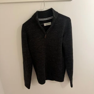 Mörkgrå stickad half zip-tröja  - Mörkgrå stickad tröja med half zip-dragkedja och hög krage. Tröjan har en slim passform och är perfekt att bära över en skjorta. Materialet är mjukt och värmande, och modellen har långa ärmar och ribbade muddar för en snygg look.