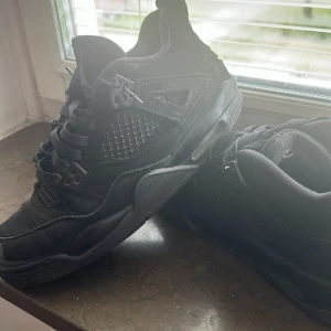 Svarta Nike Air Jordan 4 sneakers - Säljer ett par helsvarta Nike Air Jordan 4 sneakers med klassisk meshpanel, skinn och mockadetaljer. Skorna har snörning, hög siluett och ikonisk Jumpman-logga på plösen. Perfekta för dig som gillar streetstyle och vill ha en tidlös look.