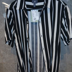 Svartvit randig skjorta från H&M - Snygg svart och vit skjorta från H&M med vertikala ränder och korta ärmar. Skjortan har klassisk krage och knäppning framtill. Perfekt för dig som gillar en stilren och trendig look. Regular fit och tillverkad i mjukt bomullstyg.
