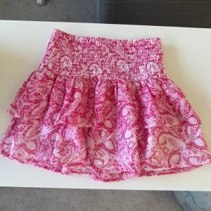 Rosa volangkjol med mönster - Supersöt kort kjol i rosa och vitt med volanger och smock i midjan. Kjolen har ett detaljerat mönster och är gjord i ett lätt och luftigt tyg, perfekt för varma dagar. Den har en feminin och lekfull vibe som passar perfekt till sneakers eller sandaler. Storlek 134/140, original pris är 229,95 sek. Ny utan prislapp. 