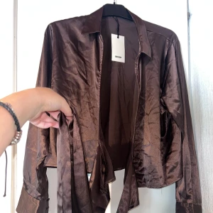 Brun satinskjorta från Bik Bok - Snygg brun skjorta i satin från Bik Bok med lång ärm och klassisk krage. Skjortan har matchande tygklädda knappar vid ärmsluten och ett bälte i midjan för att knyta till en cool look. Kan justeras till olika storlekar