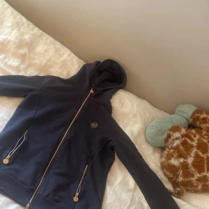 Mörkblå hoodie från Cavalleria Toscana - Snygg mörkblå hoodie från Cavalleria Toscana med guldfärgade dragkedjor och logga på bröstet. Tröjan har huva, långa ärmar och diskret mönster längs ärmarna. Perfekt för dig som gillar stilren och sportig look.