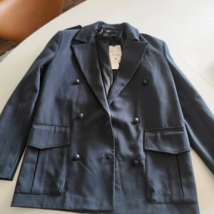 Svart kavaj från Zara, dubbelknäppt XS - Stilren svart kavaj från Zara i storlek XS. Dubbelknäppt modell med breda slag, två stora fickor framtill och knappar vid ärmslut. Perfekt för dig som vill ha en klassisk och snygg look. Materialet är slätt och känns bekvämt mot huden.