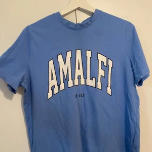 Blå t-shirt med Amalfi-tryck - Snygg blå t-shirt med stort vitt tryck där det står 'AMALFI' och mindre text 'ITALY' under. Klassisk rund hals och korta ärmar, perfekt för en chill och avslappnad stil.