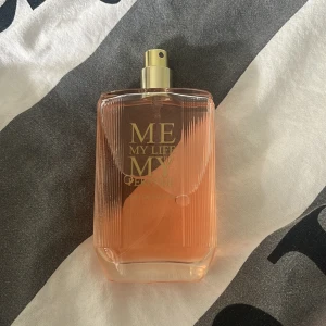 Me My Life My Perfume Eau de Parfum - En stilren parfym i en rektangulär glasflaska med räfflade kanter och guldig spraykork. Flaskan har en ljusrosa ton och texten 'ME MY LIFE MY PERFUME' i guld på framsidan. Perfekt för dig som gillar snygg design och fräscha dofter. Kostade runt 100kr helt ny pris går att diskutera!