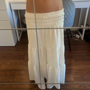 Vit långkjol med smock i midjan - Superfin vit långkjol med smockad midja och volangiga lager. Kjolen har en lätt skrynklig struktur och . Passar dig som gillar flowiga och bekväma plagg.har en smutfäck bak men syns inte så bra 