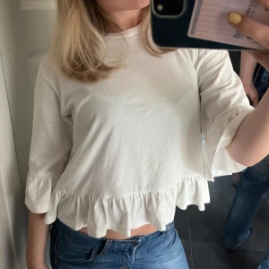 Vit volangblus med trekvartsärm - Supersöt vit blus med volangdetaljer längs nederkanten och ärmsluten. Modellen är croppad och har trekvartsärmar som ger en avslappnad men ändå trendig look. Perfekt att matcha med jeans för en chill stil.