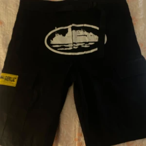Svarta cargo shorts Corteiz Guerillaz - Svarta cargo shorts från Corteiz Guerillaz med vita tryckdetaljer och broderad logga. Stora fickor på sidorna och bak, samt gul patch med text. Klassisk loose fit och robust känsla, perfekta för streetwear. Materialet känns som bomull eller polyester.