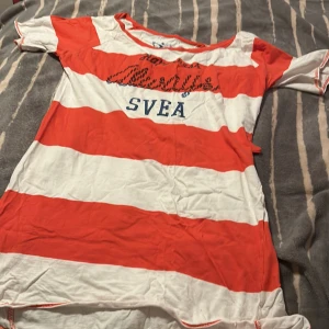 Randig rödvit klänning från Svea XS - Säljer en kort klänning från Svea i storlek XS med breda röda och vita ränder. Klänningen har rund halsringning, korta ärmar och tryckt Svea-logga framtill. Tillverkad i mjuk bomull, perfekt för en avslappnad och sportig look. Pyjamas 