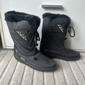 Svarta vinterboots med fuskpäls - Svarta boots med mjuk fuskpäls runt skaftet och snörning framtill. Snygga detaljer med broderade mönster i beige och svart. Sulan är platt och grov för bra grepp. Perfekta för kalla dagar och ger en cool vinterlook.