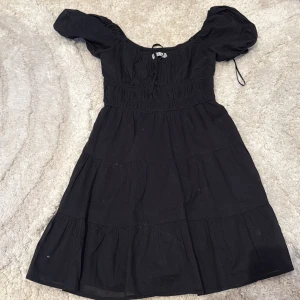 Svart kortklänning från Hollister XS - Supersöt svart klänning från Hollister i bomull med puffärmar och smockad midja. Klänningen har knytning vid halsen och volangiga lager i kjolen. Perfekt för dig som gillar romantisk och avslappnad stil.