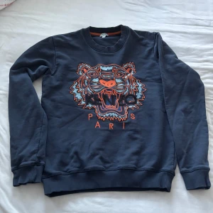Mörkblå Kenzo sweatshirt med tiger - Säljer en mörkblå sweatshirt från Kenzo med ikoniskt broderat tigerhuvud och Paris-text i orange och blått framtill. Tröjan har rund hals, ribbade muddar och är långärmad. Perfekt statement piece för dig som gillar streetstyle. Äkta!