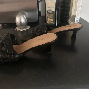Svarta flätade sandaler med klack - Stilrena svarta sandaler från Amezy med låg klack och fyrkantig tå. Ovandelen har en bred, flätad rem i skinnimitation som ger en trendig look. Perfekta för dig som gillar en chic och modern stil.