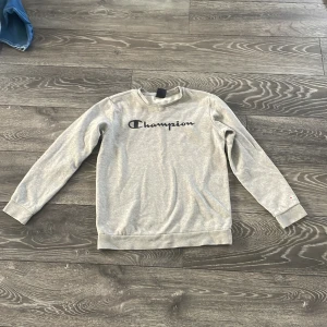 Grå Champion sweatshirt X-Large - Snygg grå sweatshirt från Champion i storlek X-Large (162/167). Tröjan har svart Champion-logga på bröstet och en liten logga på ärmen. Klassisk crewneck med långa ärmar och mjukt material, perfekt för chill dagar.