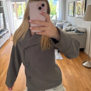 Grå kappa - Säljer en stilren grå kappa med stora knappar framtill. Jackan har en rak och lite boxig passform, perfekt för dig som gillar en klassisk men ändå trendig look. Materialet är mjuk ull som håller dig varm under kyliga dagar.